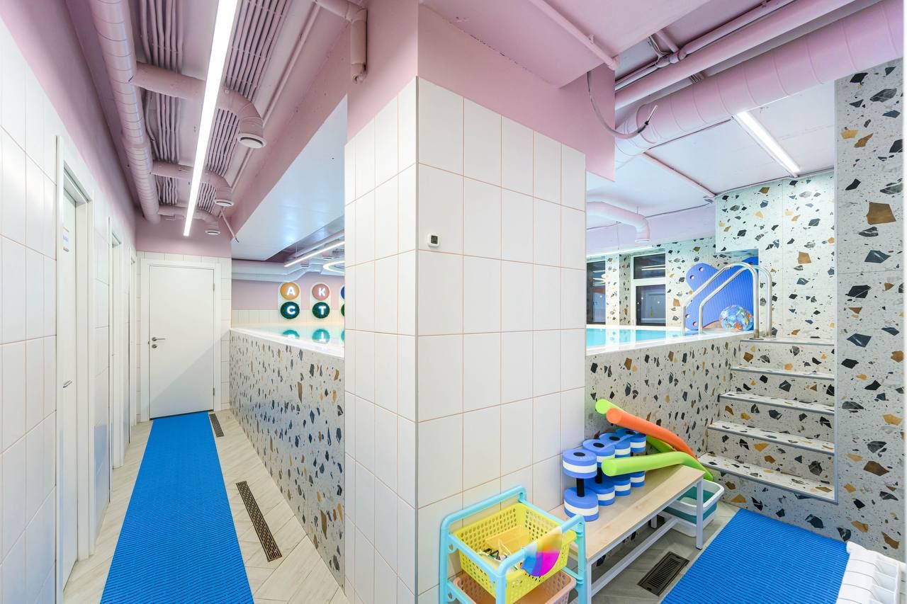 Colorful indoor pool area with a tiled wall, blue mats, and a basket of pool toys. Stairs lead to a pool with patterned walls and playful decor.
