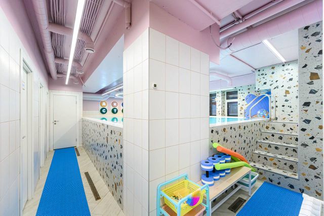 Colorful indoor pool area with a tiled wall, blue mats, and a basket of pool toys. Stairs lead to a pool with patterned walls and playful decor.