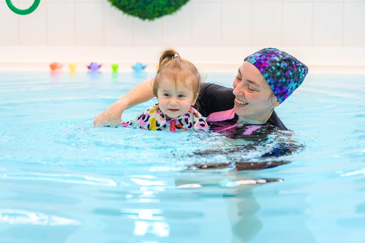 Adult guiding a toddler in a swimming pool, both smiling. Colorful floating toys are visible in the background.