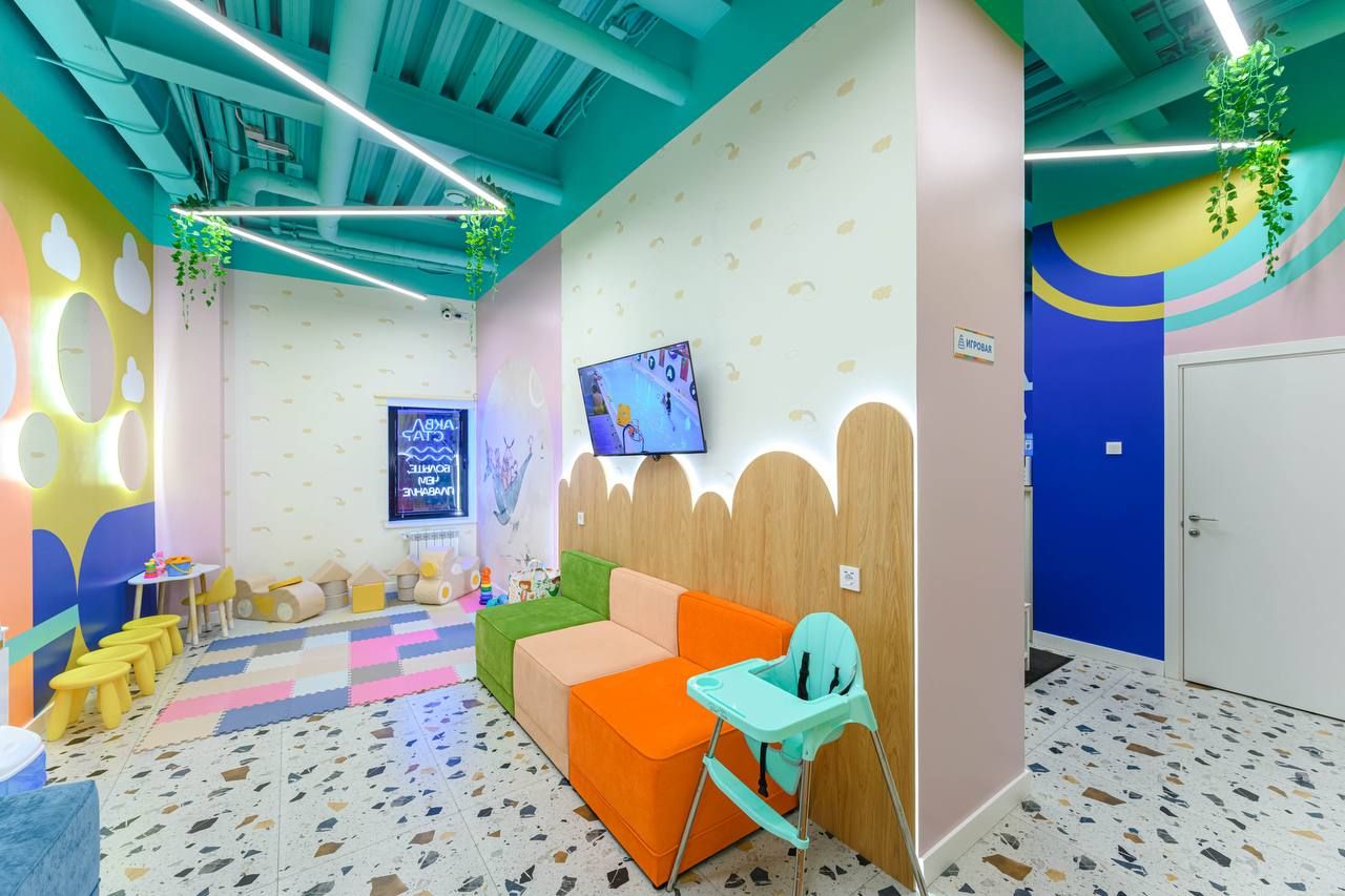 Bright children's playroom with colorful walls, a TV, seating, a high chair, and a play area with small tables and chairs.