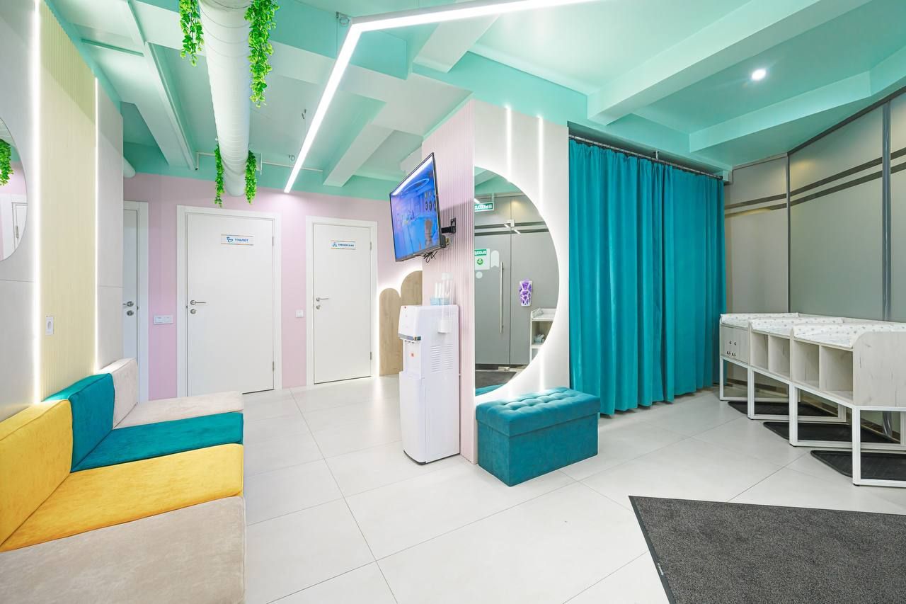 Modern waiting area with colorful seating, a TV, water cooler, and teal curtains. Bright lighting and plants add a fresh touch.