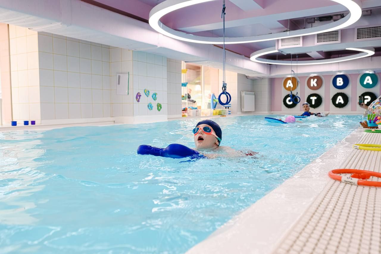 Child swimming in an indoor pool wearing goggles and a swim cap. Brightly lit space with colorful decorations and large circular lights overhead.