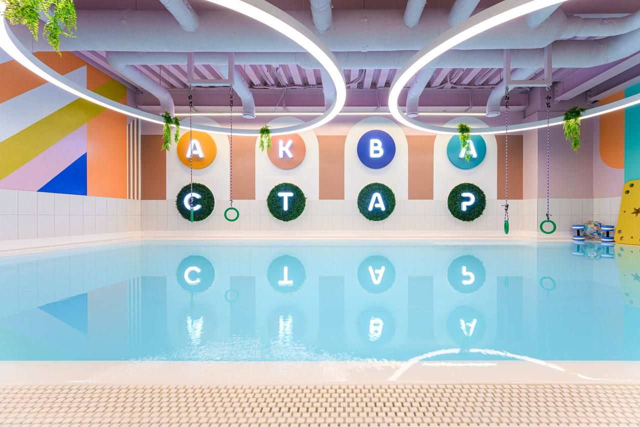 Indoor pool with colorful walls, circular light fixtures, and decorative greenery. Large letters on the wall reflect in the water.