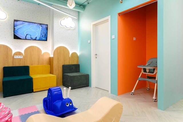 Colorful children's playroom with a TV, cloud wall decor, colorful seating, a blue rocking snail, and a high chair in an orange alcove.