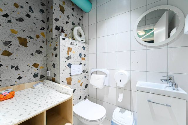Modern bathroom with a changing table, toilet, sink, and round mirror. Walls have a mix of white tiles and terrazzo patterns.