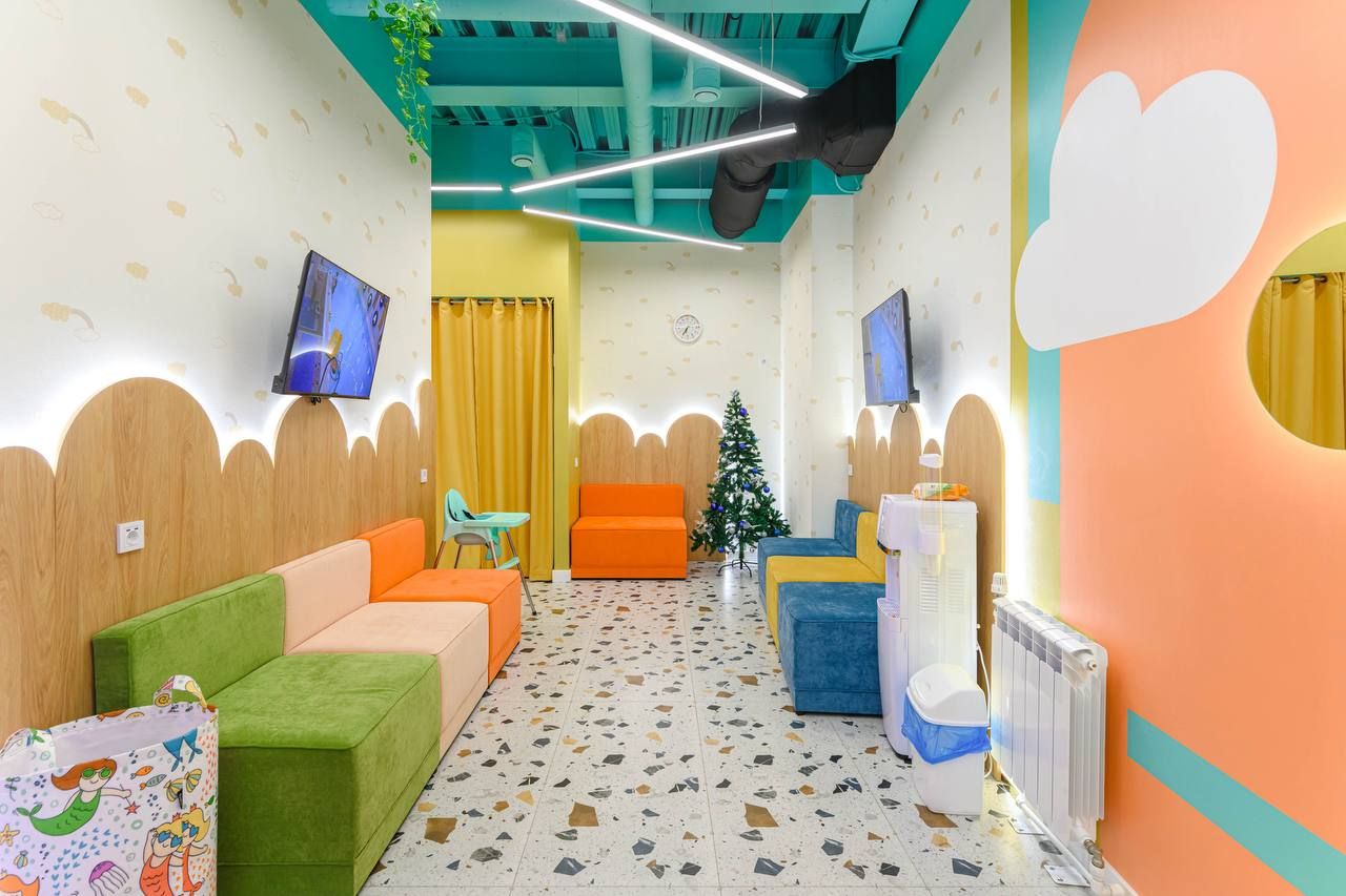 Bright playroom with colorful seating, wall murals, a small Christmas tree, and two TVs. Terrazzo floor and playful decor create a cheerful atmosphere.