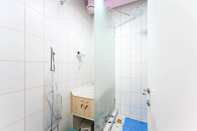 A small bathroom with white tiled walls, a glass shower, a wall-mounted hairdryer, and a wooden cabinet with a patterned surface.