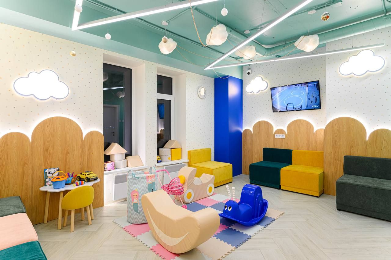 Bright children's playroom with colorful seating, toys, cloud-shaped lights, and a TV on the wall. Playful and inviting atmosphere.