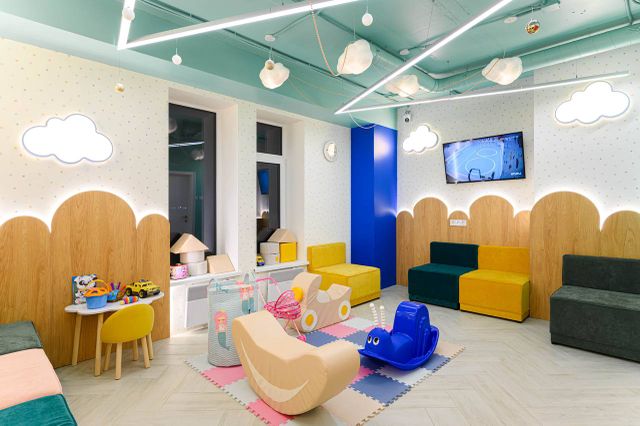 Bright children's playroom with colorful seating, toys, cloud-shaped lights, and a TV on the wall. Playful and inviting atmosphere.