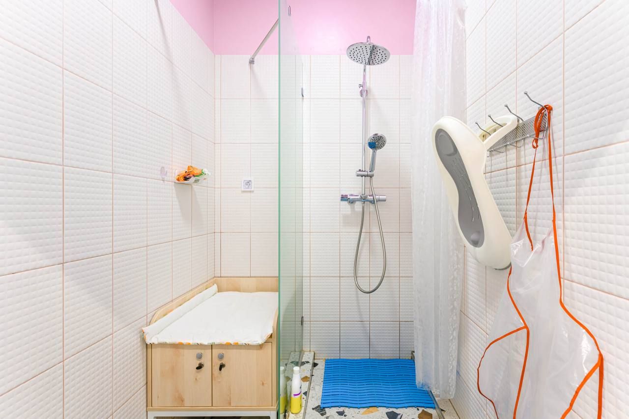 A small bathroom with a shower, glass partition, changing table, blue mat, and wall hooks. Pink ceiling contrasts with white tiled walls.
