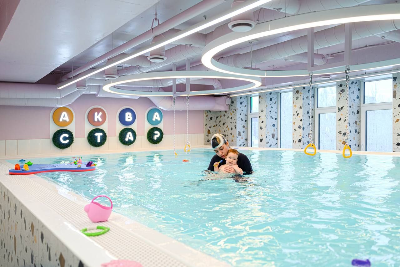 A parent and child swim in an indoor pool with colorful toys. Bright circular lights hang above, and large windows line the wall.