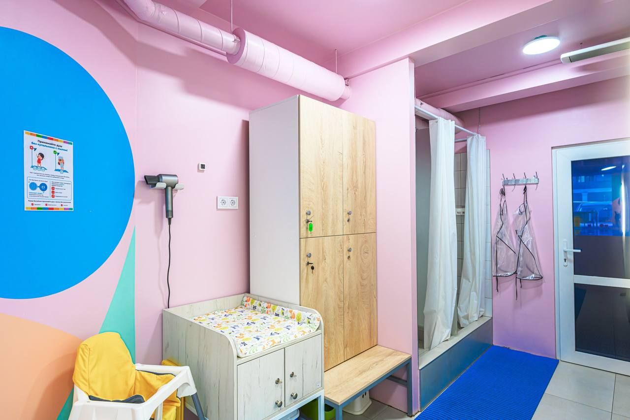 Brightly colored baby changing room with pink walls, wooden cabinets, a changing table, high chair, and informational poster.