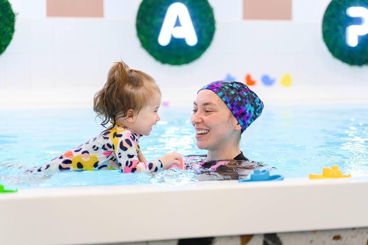 A smiling adult and child in colorful swimwear enjoy a swim in a pool, surrounded by floating toys and decorative letters on the wall.