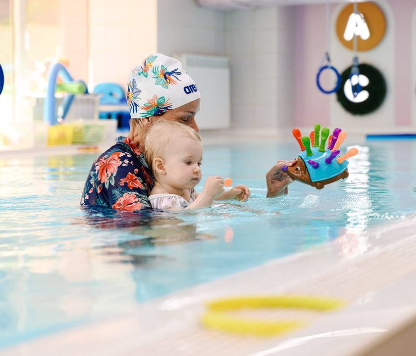 An adult and a toddler in a swimming pool, playing with colorful toys. The adult wears a floral swimsuit and a swim cap.
