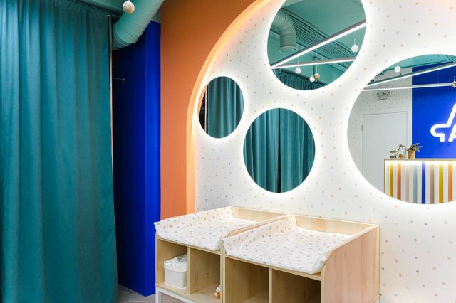 Brightly lit modern changing room with two wooden changing tables, circular mirrors on a polka dot wall, and teal curtains.