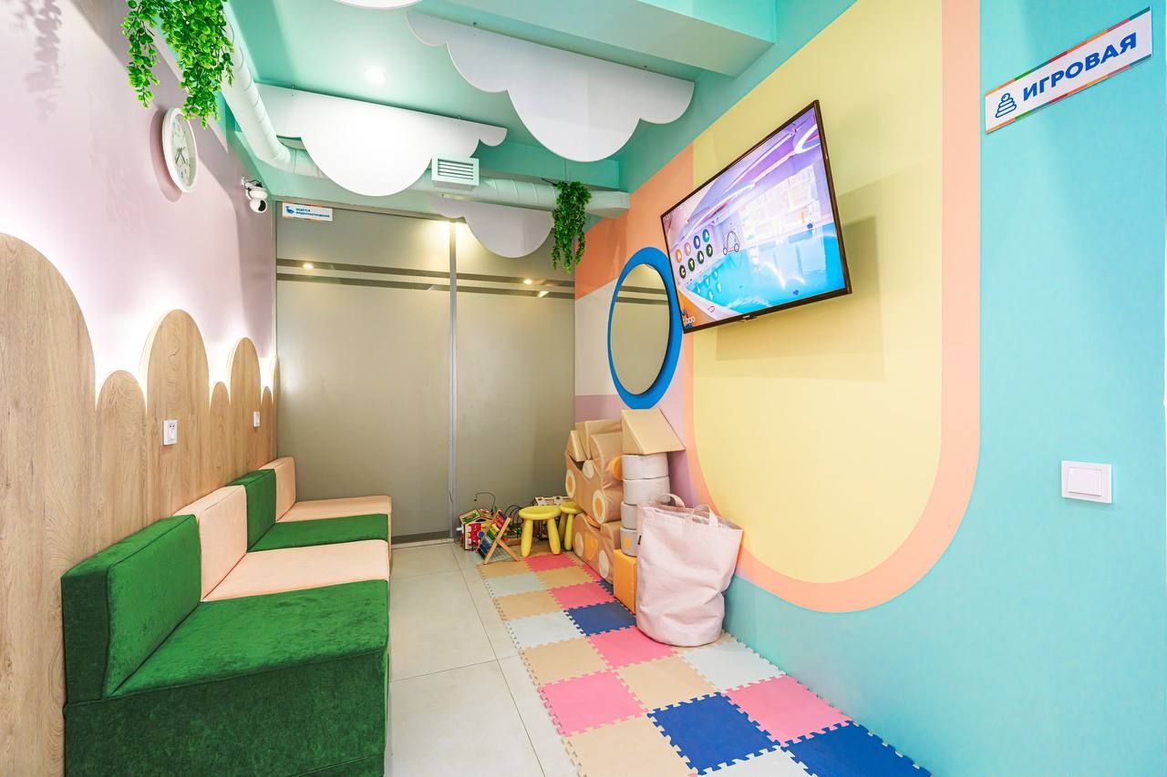Colorful children's playroom with pastel walls, green seating, a TV, toys, and foam mat flooring. Ceiling features cloud decorations and plants.