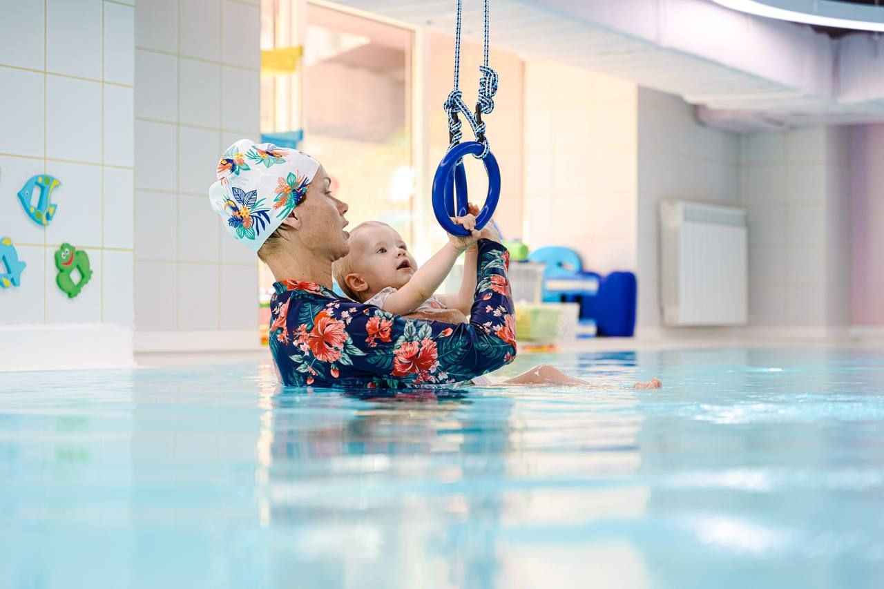 Adult and baby in a swimming pool, both wearing colorful swim caps. The baby reaches for a blue ring hanging above the water.