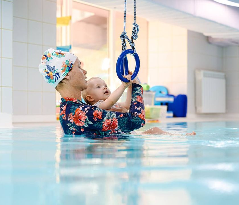 Adult and baby in a swimming pool, both wearing colorful swim caps. The baby reaches for a blue ring hanging above the water.