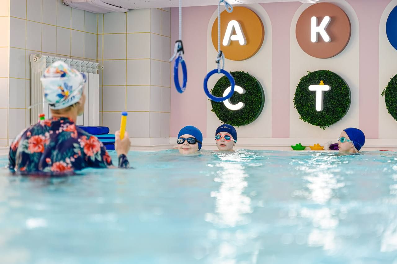 Swimming instructor with three children in a pool, all wearing swim caps and goggles. Wall has colorful letters and decorative greenery.