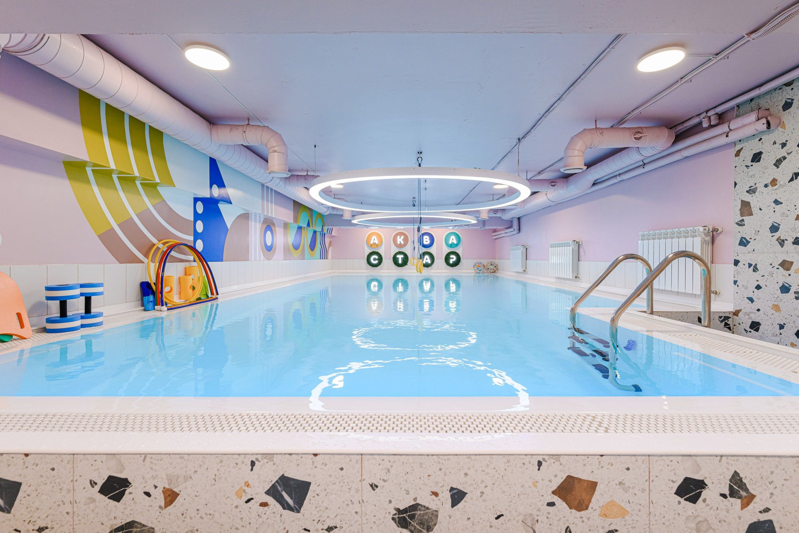 Indoor swimming pool with colorful wall art, ring-shaped light fixture, and pool toys. Steps lead into the clear blue water.