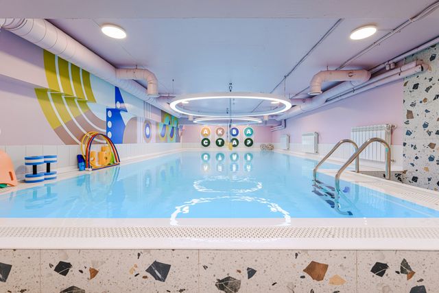 Indoor swimming pool with colorful wall art, ring-shaped light fixture, and pool toys. Steps lead into the clear blue water.