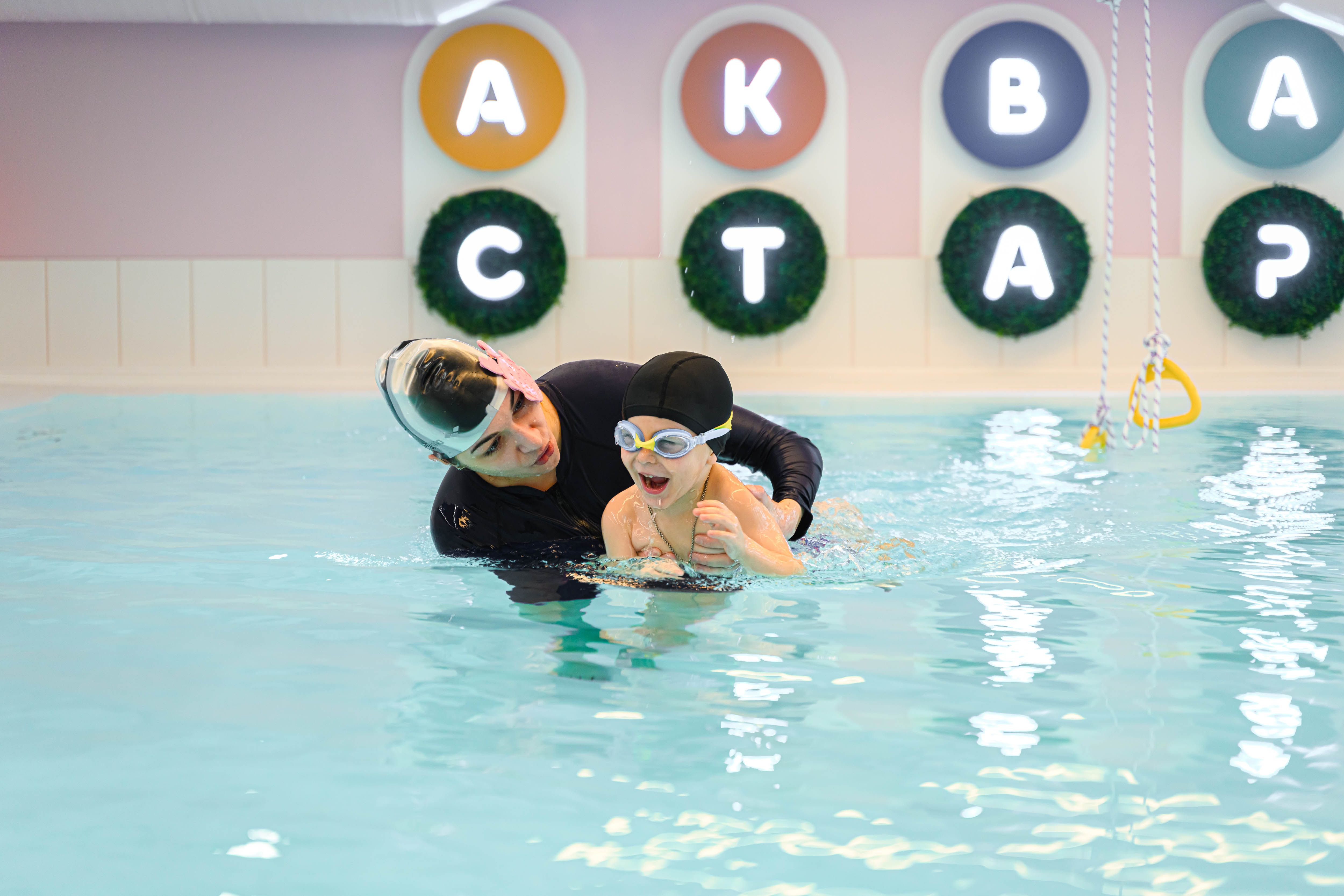 Instructor helping a child learn to swim in a pool, both wearing swim caps and goggles, with colorful letters on the wall in the background.