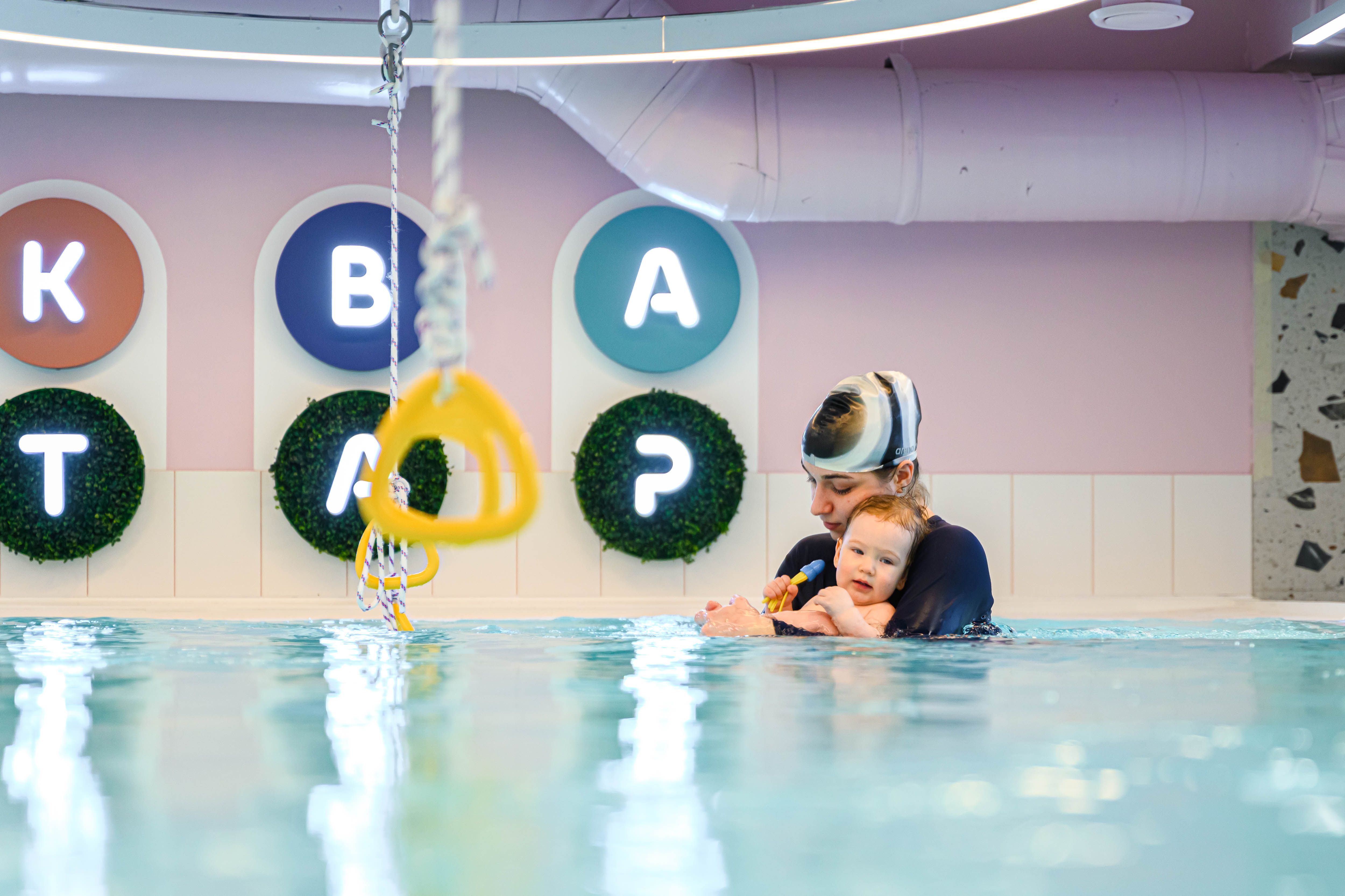 Instructor holding a toddler in a swimming pool, with colorful wall decorations in the background.
